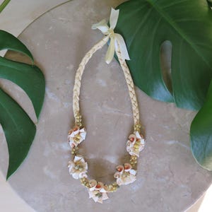May include: A handcrafted necklace featuring a braided straw design with seashell accents. The necklace has a cream-colored ribbon tie and is adorned with various seashells and small beads. The necklace is displayed on a marble surface with green leaves.