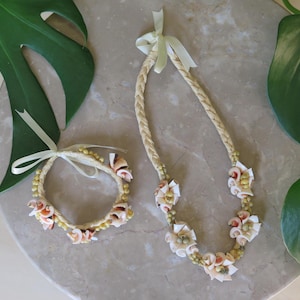 May include: A matching jewelry set featuring a necklace and bracelet. The necklace and bracelet are made of woven beige material, adorned with seashells and small yellow beads. Both pieces are tied with a light green ribbon.