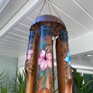 Op de afbeelding: Een windgong met een donkerbruine kokosnootschaal aan de bovenkant en verschillende holle bamboebuizen. De buizen zijn versierd met handgeschilderde roze en blauwe hibiscusbloemen en een bruin rasterpatroon. Het hangt aan een zwart koord.