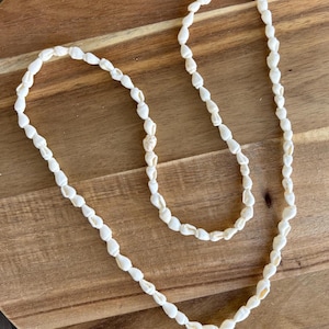 Puede incluir: Un collar hecho de pequeñas conchas blancas. Las conchas están ensartadas, formando una larga hebra en bucle. El collar se exhibe sobre una superficie de madera, mostrando su estética natural y orgánica. Las conchas tienen un color blanco cremoso.