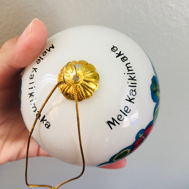 May include: White Christmas ornament with a gold top and a gold string. The ornament has the text "Mele Kalikimaka" written on it in black.