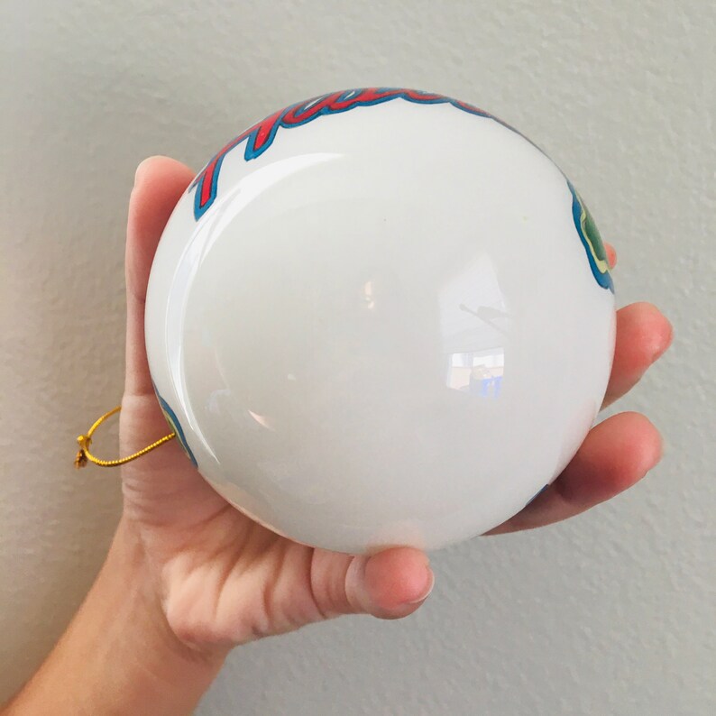 May include: White Christmas ornament with a blue, red, and green design. The ornament has the word "Hawaii" written on it.