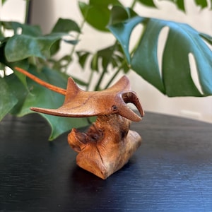 Op de afbeelding: Een handgemaakte houten sculptuur van een manta rog, met een gedetailleerd ontwerp en een natuurlijke houtnerf afwerking. De sculptuur is gemonteerd op een stuk getextureerd hout, wat de vaardigheid van de kunstenaar laat zien. De achtergrond bevat groen gebladerte.