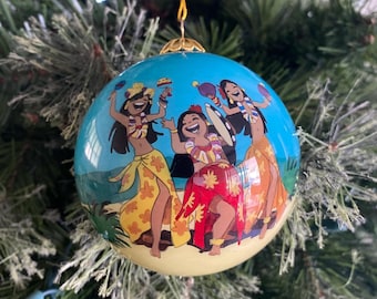 Puede incluir: Un adorno navideño redondo azul con una escena de playa con tres mujeres con vestidos coloridos y collares de flores tocando instrumentos musicales. El adorno tiene una cuerda dorada para colgar.