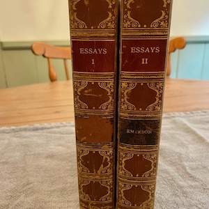 K&ouml;nnte beinhalten: Zwei antike, ledergebundene B&uuml;cher mit goldenen Verzierungen und kunstvollen Designs. Die B&uuml;cher tragen die Titel "Essays I" und "Essays II" und der Name des Autors "Emerson" steht auf dem zweiten Buch.