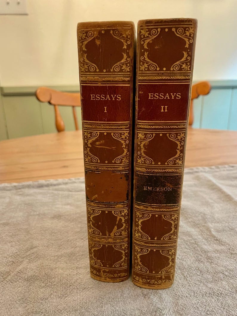 K&ouml;nnte beinhalten: Zwei antike, ledergebundene B&uuml;cher mit goldenen Verzierungen und kunstvollen Designs. Die B&uuml;cher tragen die Titel "Essays I" und "Essays II" und der Name des Autors "Emerson" steht auf dem zweiten Buch.