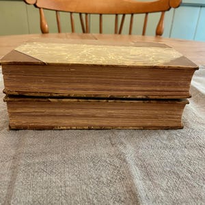 K&ouml;nnte beinhalten: Zwei antike B&uuml;cher mit braunen Ledereinb&auml;nden und goldfarbenem Marmorpapier auf der Vorderseite. Die B&uuml;cher liegen &uuml;bereinander auf einer braunen, strukturierten Oberfl&auml;che.