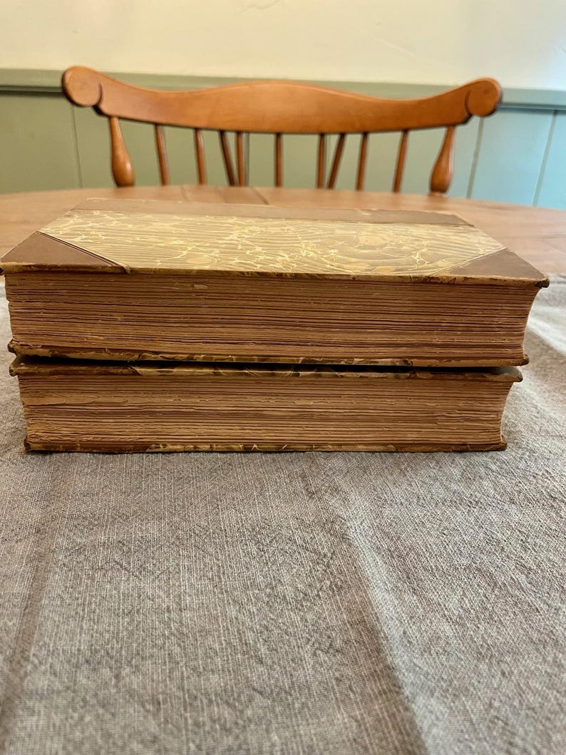 K&ouml;nnte beinhalten: Zwei antike B&uuml;cher mit braunen Ledereinb&auml;nden und goldfarbenem Marmorpapier auf der Vorderseite. Die B&uuml;cher liegen &uuml;bereinander auf einer braunen, strukturierten Oberfl&auml;che.