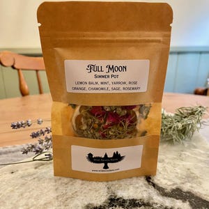 May include: A brown paper pouch with a white label that reads "FULL MOON SIMMER POT LEMON BALM, MINT, YARROW, ROSE ORANGE, CHAMOMILE, SAGE, ROSEMARY" and a smaller label that reads "Wild Raven Farm www.wildravenfarm.com". The pouch contains a blend of dried herbs and flowers.