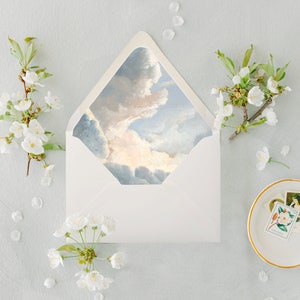 May include: A white envelope with a sky blue and pink painted design on the inside flap. The envelope is surrounded by white flower blossoms and petals on a light blue background.