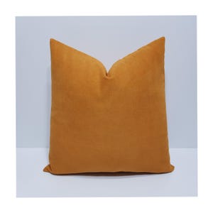 May include: A square throw pillow in a solid, deep mustard yellow color. The pillow features a soft, matte fabric texture with a distinct crease at the top center, creating a subtle indentation that gives the cushion a plump and structured appearance.