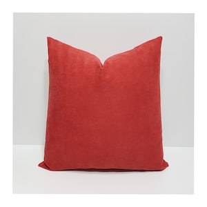May include: A square throw pillow featuring a solid, deep red suede-like fabric with a soft, matte texture. The pillow has a distinct, centered indentation at the top edge, creating a classic karate-chop shape that gives it a structured appearance.