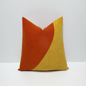 Può includere: Un cuscino quadrato caratterizzato da un design bicolore con una cucitura diagonale. Il lato sinistro è di un arancione bruciato intenso, mentre il lato destro è di un giallo senape vivace. Il tessuto presenta una consistenza morbida e opaca.
