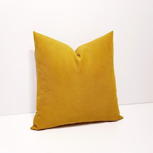 May include: A square throw pillow in a solid mustard yellow color. The pillow features a soft, matte fabric texture with a clean, simple design and a visible seam along the edges, presenting a uniform appearance without any patterns or prints.