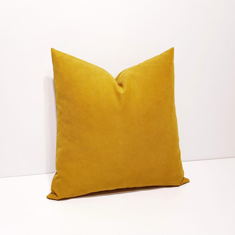 May include: A square throw pillow in a solid mustard yellow color. The pillow features a soft, matte fabric texture with a clean, simple design and a visible seam along the edges, presenting a uniform appearance without any patterns or prints.