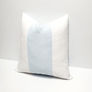 May include: A square throw pillow featuring a solid white fabric on the left and right sides, with a wide, vertical, light blue panel centered across the middle. The pillow has a smooth, matte texture and clean, straight seams along the color blocks.