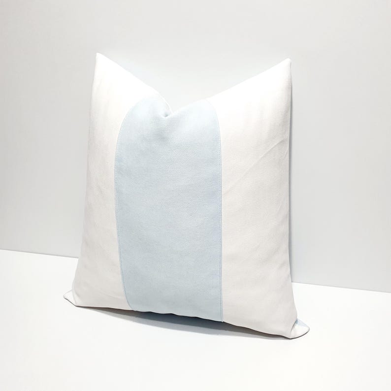 May include: A square throw pillow featuring a solid white fabric on the left and right sides, with a wide, vertical, light blue panel centered across the middle. The pillow has a smooth, matte texture and clean, straight seams along the color blocks.