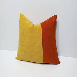 May include: A square throw pillow featuring a color-blocked design with a large section of mustard yellow fabric on the left and a smaller vertical section of burnt orange fabric on the right, both made of a soft, matte, velvet-like textile material.
