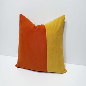 May include: A square throw pillow featuring a color-blocked design with a deep orange section on the left and a vibrant mustard yellow section on the right. The fabric has a soft, velvet-like texture with clean, straight vertical seams.