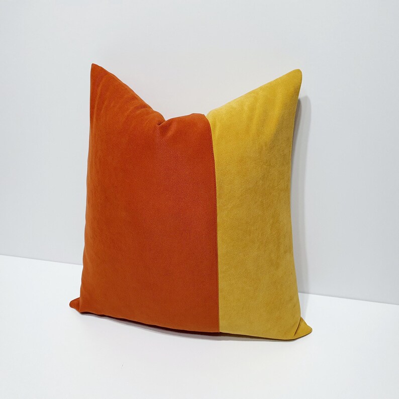 May include: A square throw pillow featuring a color-blocked design with a deep orange section on the left and a vibrant mustard yellow section on the right. The fabric has a soft, velvet-like texture with clean, straight vertical seams.