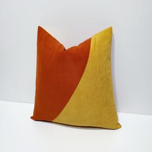 May include: A square cushion with a two-tone design. One half is a burnt orange colour, and the other half is a golden yellow. The cushion is made of a soft, textured fabric and is likely used for home decor.