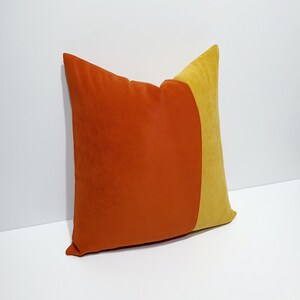 May include: A square throw pillow featuring a color-blocked design with a large section of burnt orange velvet fabric on the left and a smaller section of mustard yellow velvet fabric on the right, creating a distinct vertical division across the surface.