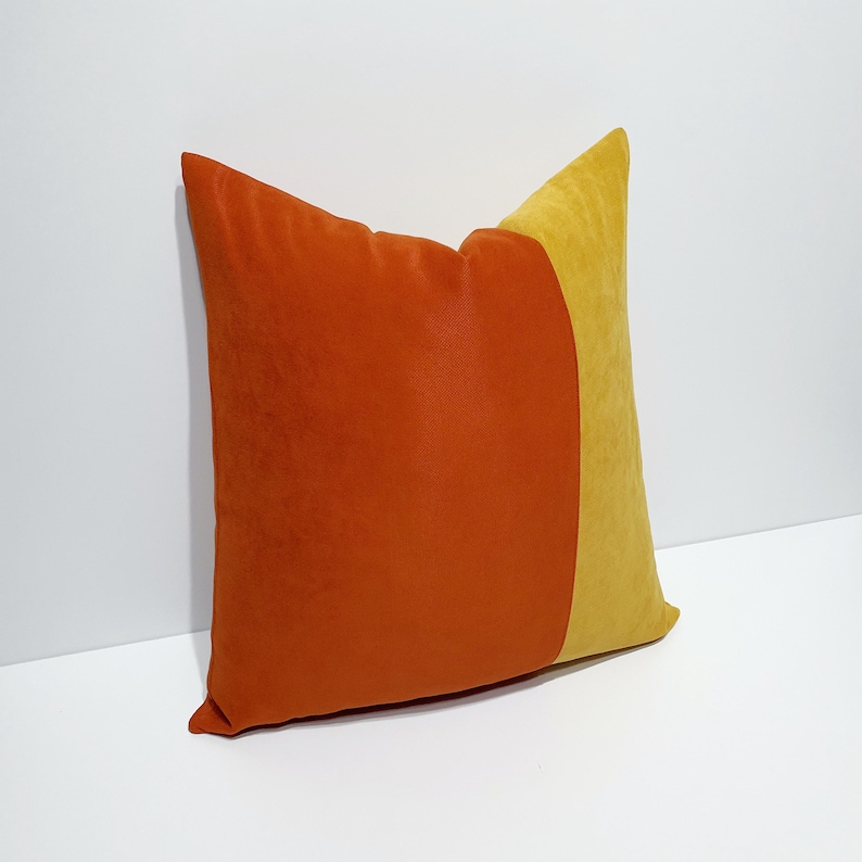 May include: A square throw pillow featuring a color-blocked design with a large section of burnt orange velvet fabric on the left and a smaller section of mustard yellow velvet fabric on the right, creating a distinct vertical division across the surface.