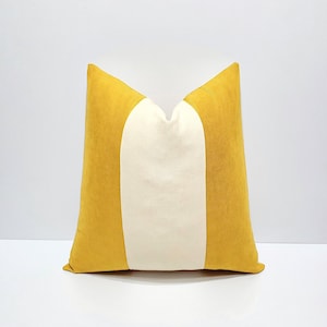 Pode incluir: Uma almofada quadrada com um design de blocos de cor, apresentando um painel central vertical largo na cor creme, ladeado por dois painéis laterais em amarelo mostarda sólido. O tecido tem uma textura macia e mate, com formato pontiagudo.