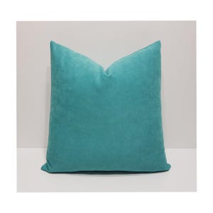May include: A square throw pillow featuring a solid teal color with a soft, matte, suede-like texture. The pillow has a clean, minimalist design with visible seams along the edges, presenting a uniform and consistent appearance throughout the surface.