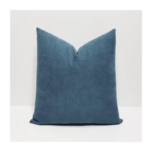 Puede incluir: Un cojín cuadrado de color azul cerceta profundo y sólido. La tela tiene una textura suave, mate y similar a la gamuza con un acabado de grano fino visible. El cojín está ahuecado en el centro superior, creando una hendidura en forma de V.