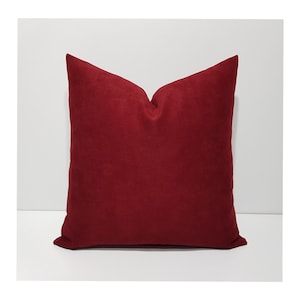 May include: A square throw pillow featuring a solid deep red color with a soft, suede-like texture. The pillow has a distinct, plump shape with a slight indentation at the top center, creating a clean, uniform appearance across the entire surface.