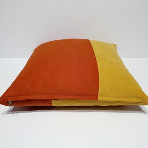 May include: A square throw pillow featuring a color-blocked design with a large section of burnt orange on the left and a smaller section of mustard yellow on the right. The pillow has a visible silver zipper closure along the bottom edge.