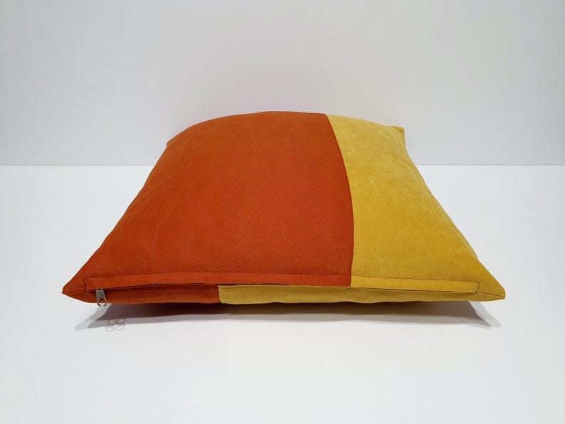 May include: A square throw pillow featuring a color-blocked design with a large section of burnt orange on the left and a smaller section of mustard yellow on the right. The pillow has a visible silver zipper closure along the bottom edge.
