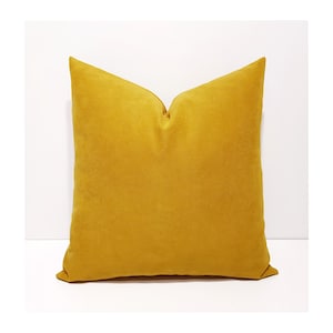 May include: A square scatter cushion in a solid mustard yellow colour. The fabric has a soft, matte texture with a visible weave. The cushion is plumped to create a slight indentation at the top centre, giving it a structured and full appearance.