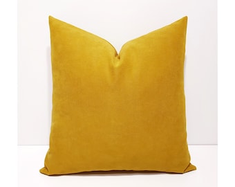 May include: A square throw pillow in a solid mustard yellow color. The fabric has a soft, matte texture with a visible weave. The pillow is plumped to create a slight indentation at the top center, giving it a structured and full appearance.