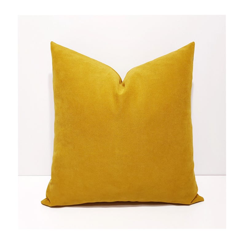 May include: A square throw pillow in a solid mustard yellow color. The fabric has a soft, matte texture with a visible weave. The pillow is plumped to create a slight indentation at the top center, giving it a structured and full appearance.