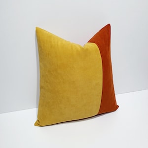 May include: A square throw pillow featuring a color-blocked design with a large mustard yellow section on the left and a smaller burnt orange section on the right. The fabric has a soft, matte, suede-like texture throughout the entire surface.