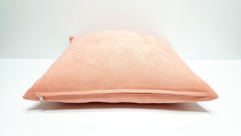 May include: A square throw pillow cover in a solid light peach color. The fabric has a soft, matte texture and features a visible metal zipper closure along the bottom edge, allowing for easy removal and insertion of a pillow insert.