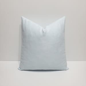 May include: A square throw pillow in a solid, light powder blue color. The pillow features a smooth, uniform fabric texture with clean, straight edges and a slightly plump, filled appearance, creating a simple and consistent geometric shape.