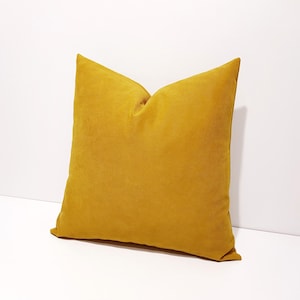 May include: A square throw pillow in a solid mustard yellow color. The pillow features a soft, matte fabric texture with a visible seam along the edges, creating a structured shape with a slight indentation at the top center of the cushion.