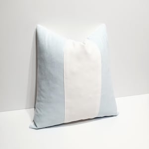 May include: A square throw pillow featuring a vertical color-blocked design. The pillow has a wide, solid white center panel flanked by two light blue side panels. The fabric appears to have a soft, smooth texture with clean, straight seams.