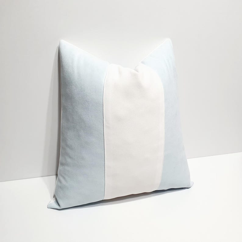 May include: A square throw pillow featuring a vertical color-blocked design. The pillow has a wide, solid white center panel flanked by two light blue side panels. The fabric appears to have a soft, smooth texture with clean, straight seams.