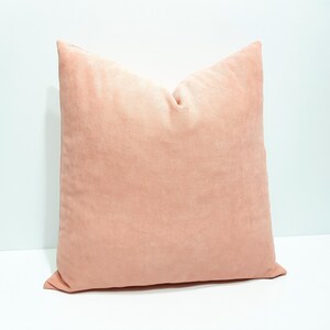 May include: A square throw pillow in a solid, muted peach color. The fabric has a soft, matte texture with a visible fine-grain weave. The pillow is plumped and features clean, straight stitched edges along the perimeter of the square shape.