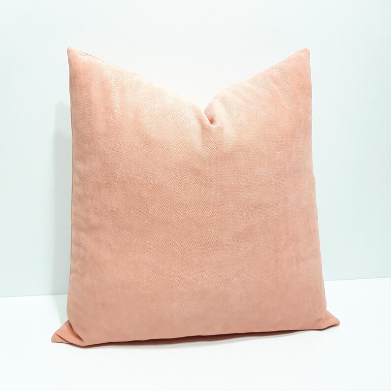 May include: A square throw pillow in a solid, muted peach color. The fabric has a soft, matte texture with a visible fine-grain weave. The pillow is plumped and features clean, straight stitched edges along the perimeter of the square shape.