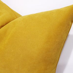 May include: A mustard yellow throw pillow featuring a distinct woven fabric texture. The square cushion displays a consistent golden-yellow hue across its surface, with visible stitching along the corner edge and a structured, plump appearance.