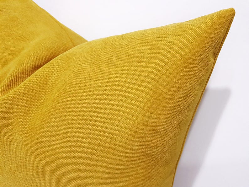 May include: A mustard yellow throw pillow featuring a distinct woven fabric texture. The square cushion displays a consistent golden-yellow hue across its surface, with visible stitching along the corner edge and a structured, plump appearance.