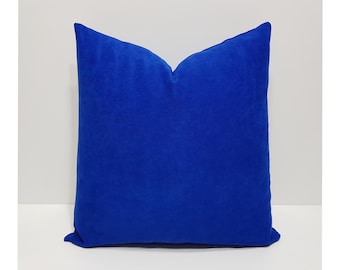 May include: A square throw cushion featuring a solid, vibrant royal blue velvet fabric. The cushion has a soft, plush texture with a distinct indentation at the top centre, creating a classic, plump shape with clean, finished edges on all sides.