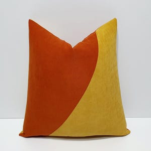 May include: A square cushion with a contemporary design. It is divided diagonally, with one half in a burnt orange hue and the other in a mustard yellow. The cushion is made of a soft material, creating a visually appealing contrast.