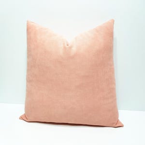 May include: A square throw pillow with a solid light peach-colored velvet cover. The fabric has a soft, matte texture and the pillow is plumped to create a slight indentation at the top center, giving it a structured, full appearance.