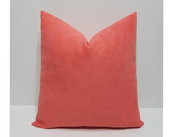 May include: A square throw pillow featuring a solid coral-colored velvet fabric cover. The pillow has a soft, matte texture and is shaped with a distinct peak at the top center, showcasing a uniform, vibrant coral hue across the entire surface.